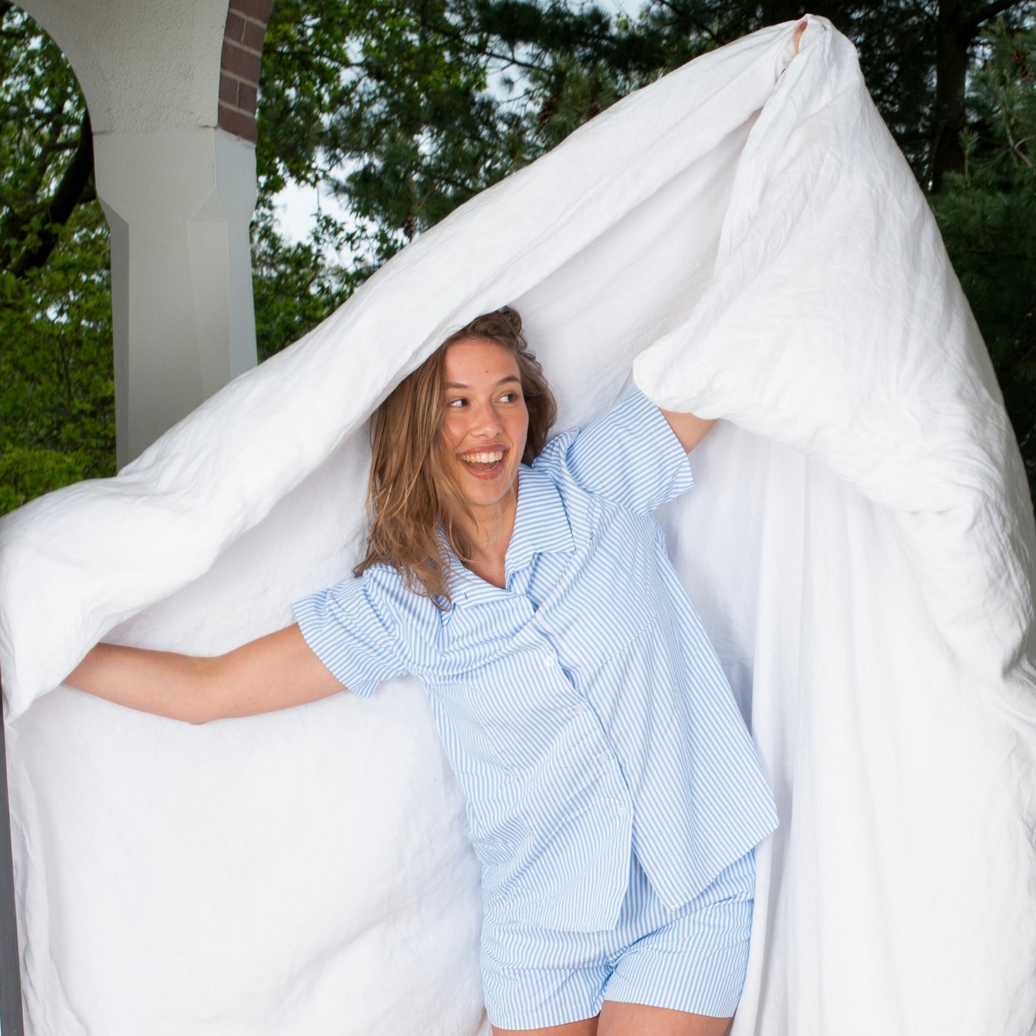 Een lachende vrouw in een blauw-wit gestreepte pyjama staat buiten onder een witte deken, met groene bomen en een deel van een zuil zichtbaar op de achtergrond. Ze kijkt opzij en ziet er vrolijk en speels uit. Katoen satijn dekbedovertrek met luxe glans -
Linnen dekbedovertrek in natuurlijke kleuren – ademend en comfortabel voor alle seizoenen