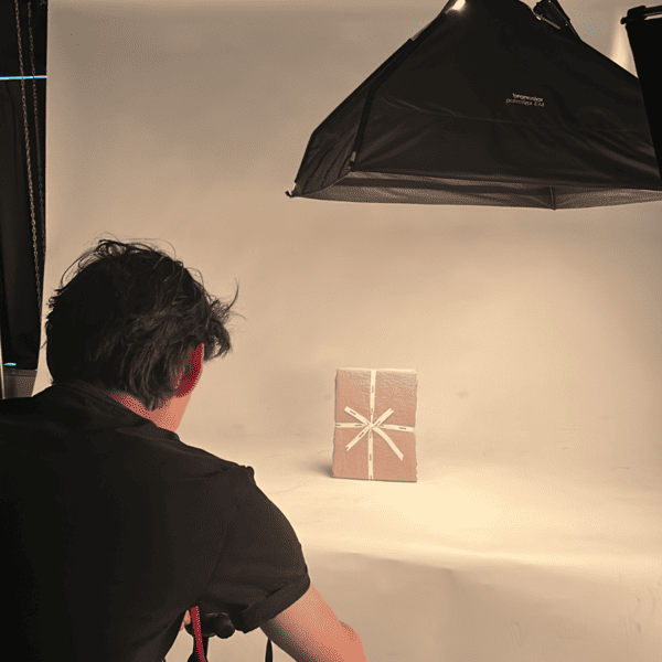 Een persoon zit in een fotostudio en kijkt naar een lichtbruin ingepakt pakket met een wit touwtje eraan vast, geplaatst op een effen achtergrond en belicht door een grote softbox.
