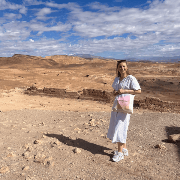 Een vrouw in een lichte jurk en sneakers staat op rotsachtige woestijngrond met een roze en beige tas in haar handen. Achter haar strekken zich bergen en een stenen muur uit onder een gedeeltelijk bewolkte blauwe hemel. Het landschap is dor, uitgestrekt en zonnig.